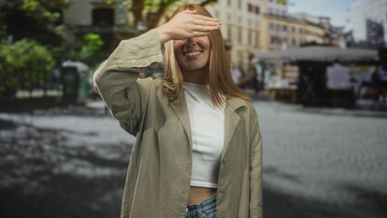 Fototapeta premium Woman covers eyes with hand while smiling, wearing cropped white top, linen jacket and jeans on a cobblestone street market in front of buildings; playful.