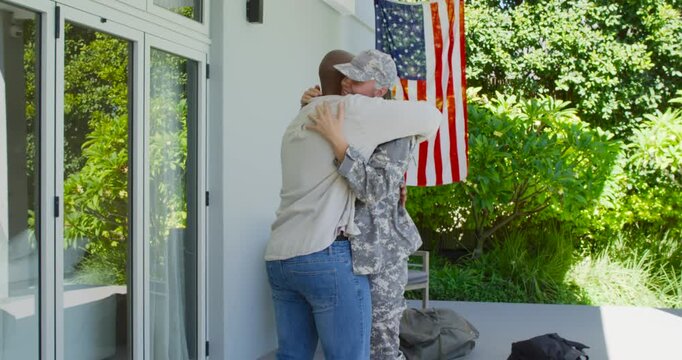 Uniformed military woman and man moving closer, embracing on porch after deployment bag and US flag