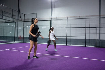 Fototapeta premium Indian and African American women in athletic dresses holding paddles, stepping on purple court