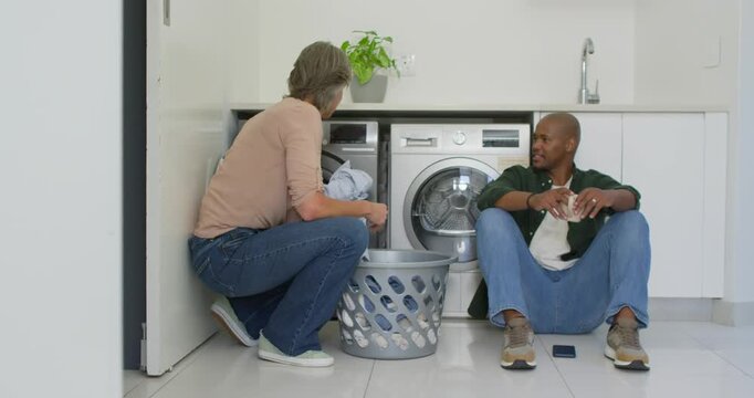 Couple sorting laundry in laundry nook, woman loading washer from basket, man watching smartphone