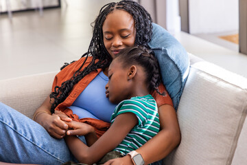Obraz premium African mother and child resting on light sofa in living room, wearing blue top and smartwatch
