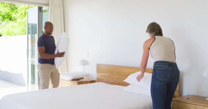 Diverse couple arranging and stacking white pillows to finish making king-size bed, smoothing duvet
