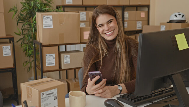 Woman holding smartphone and smiling at desk with shipping parcels and computer in a storage building; small business cheerful.