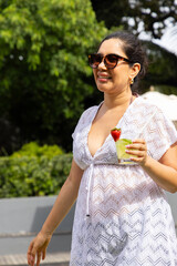 Fototapeta premium Woman walking on sunny patio wearing white lace coverup, holding mint cocktail with strawberry
