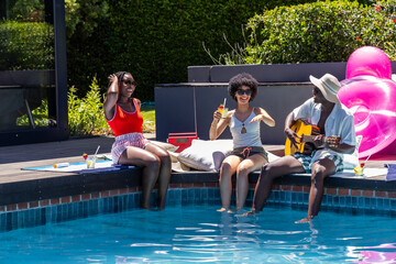 Obraz premium African American friends sitting on wooden deck at pool, in swimsuits playing guitar, holding drink