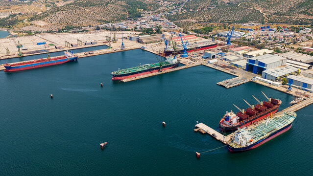 Aerial view showing large cargo ships, tankers, and bulk carriers docked at an industrial port with shipyard facilities, cranes, and vast dry docks for repair and maintenance operations