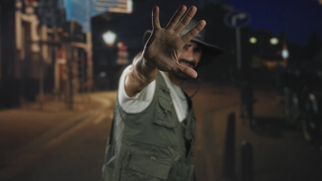 Man fisherman with beard and bucket hat extends palm facing camera on a dim street at night; guarded solitude.