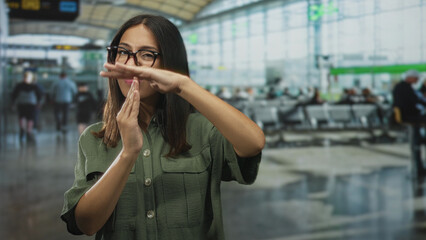 Fototapeta premium Woman with hands in timeout gesture at airport terminal wearing glasses and olive green shirt, facing camera; delay assertion.