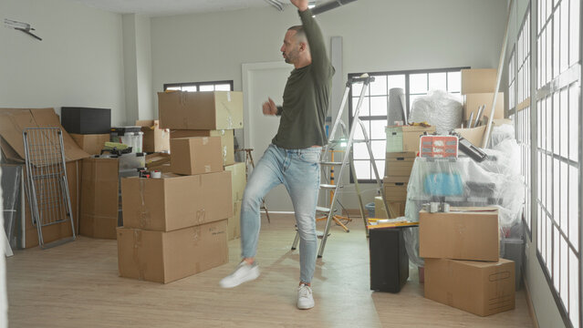 Man lifts cardboard box in a building moving scene, dancing with raised fist and stepping foot amid stacked boxes; joy fresh start.