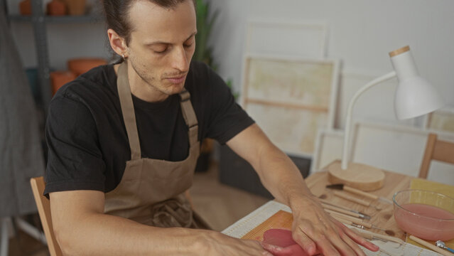 Man rolling pink clay with hands and wooden roller on a workboard in artist studio surrounded by brushes and bowls; concentration.