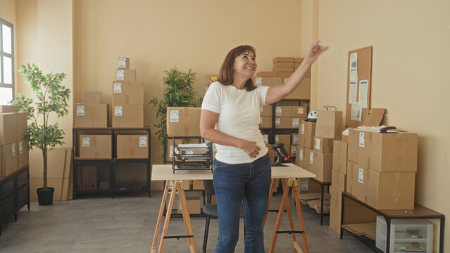 Woman pumps fists and points upward amid cardboard boxes in delivery building; small business triumph.