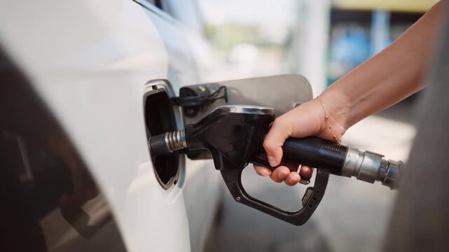 Woman refueling white car with gasoline nozzle at outdoor gas station pump bay during daytime routine
