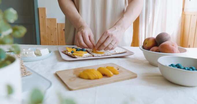 Woman making sweet sandwiches with crispbread, cream cheese, blueberry, slices of peach, coconut flakes and honey for summer picnic or brunch.