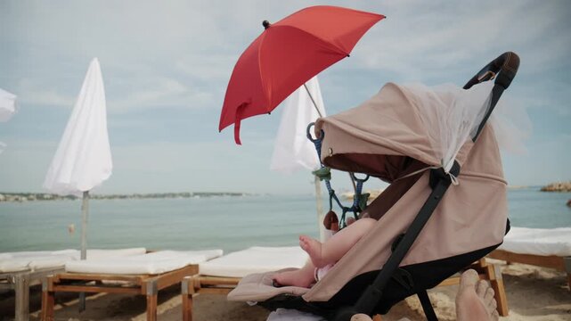 Baby sitting in stroller under bright red beach umbrella by the sea