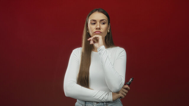 Young woman holding vaporizer, hand to chin while standing in studio with red wall and white top; pensive doubt.