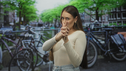 Fototapeta premium Woman forming finger gun with hands and pursed lips, visible fingers aimed forward on a street by bicycles and canal; playful confidence.