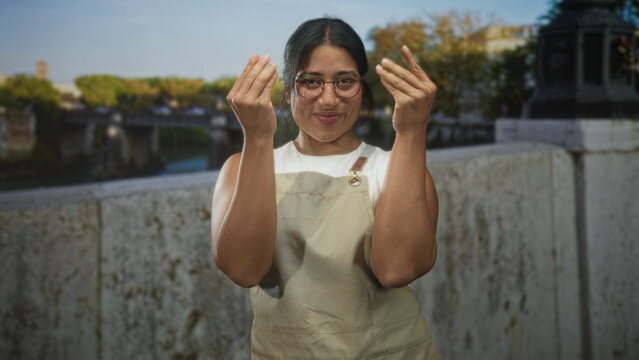 Woman in apron claps hands on street by stone bridge parapet while smiling and adjusting glasses; joy warmth friendliness.