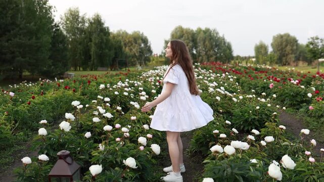 Happy teenage girl in a beautiful white summer dress spinning around and having fun in a blooming peony field with white and red flowers growing in rows on a plantation.