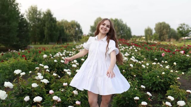 Happy teenage girl in a beautiful white summer dress spinning around and having fun in a blooming peony field with white and red flowers growing in rows on a plantation.