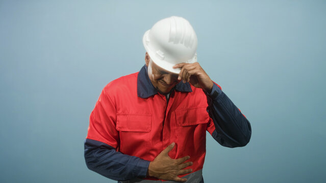 Hispanic construction worker in white hardhat and red uniform grips his stomach and tips his helmet with one hand in a studio; duty pain.