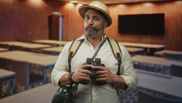 Man middle aged with beard holding binoculars, wearing backpack and pith hat in a classroom building among desks and monitor; curiosity adventure.