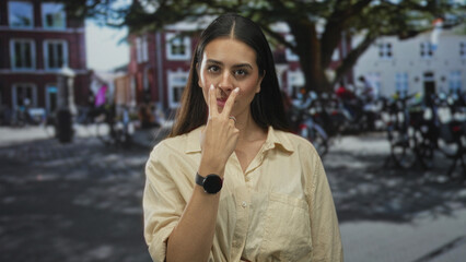 Fototapeta premium Young brunette woman points index finger in a busy street near parked motorcycles and brick buildings; firm warning.