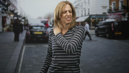 Fototapeta premium Woman clutching shoulder with hand, wincing in pain on a street lined with parked cars and shops; discomfort strain.