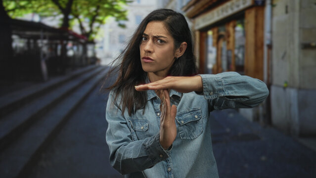 Woman makes timeout gesture with hands and palms raised in denim shirt on urban street near steps and storefronts; boundary setting frustration.