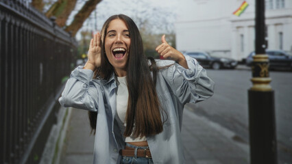Fototapeta premium Young woman cupping her hand to ear and giving a thumbs up on a city street, smiling toward camera; playful curiosity.