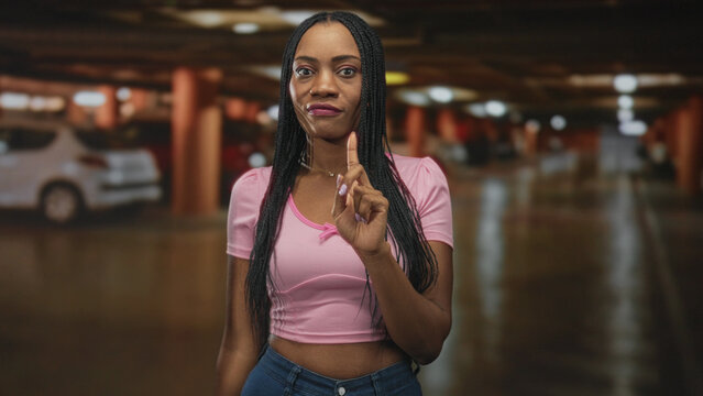 Black woman points finger forward with visible hand and pink crop top in parking building; defiance warning.