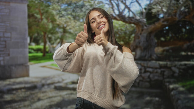 Teenage girl with braces giving double thumbs up, shoulder exposed sweater and long hair, smiling in front of stone building and leafy park path; youthful joy.