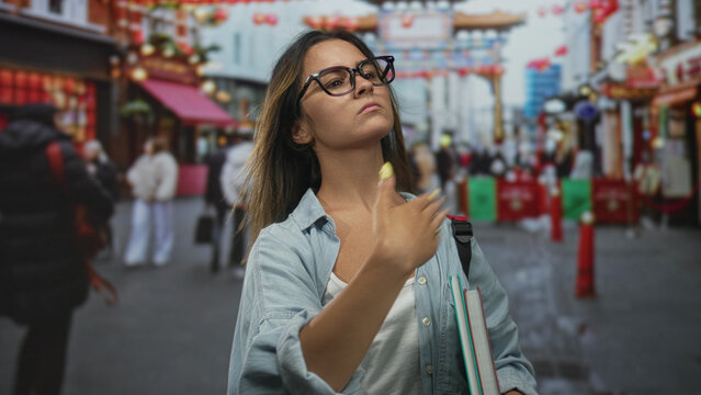 Woman touching neck while holding books and backpack on a crowded street with shops and lanterns visible; urban confidence.