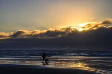 Fototapeta premium At the end of day as sun sets behind coastal cloud person walks dog on beach at water edge in silhouette