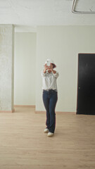 Fototapeta premium Woman architect wearing hardhat and striped shirt holding smartphone takes full length selfie in empty building room with wooden floor and column; confidence renovation.