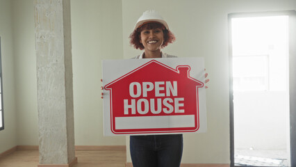 Woman holding open house sign, wearing hardhat and smiling inside the empty building; welcoming...