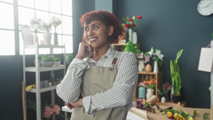 Fototapeta premium Woman florist in apron hands on hips beside bouquets and seedlings on a worktable in studio; quiet contentment.