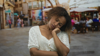 Fototapeta premium Woman with hand on cheek and eyes closed touching chin on a cobblestone street near an outdoor cafe building; pensive reflection.