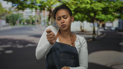 Woman dancing with fists clenched on city street wearing denim apron and long sleeve shirt, lips pursed as if singing; joy celebration.