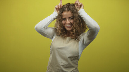 Fototapeta premium Woman in apron points fingers to head for horns gesture in studio with yellow backdrop, curly hair and warm smile; playful.
