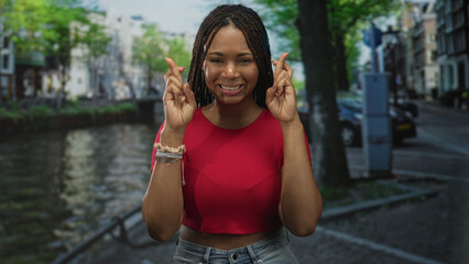 Fototapeta premium Woman clasping hands in prayer with eyes closed by amsterdam canal on a street bridge wearing red top and bracelets; hope resilience gratitude.