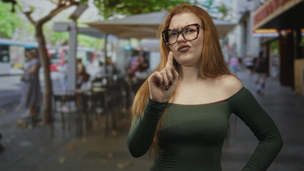 Fototapeta premium Redhead woman wearing glasses points index finger, bares shoulder in street cafe with pursed lips and raised brow; disapproval.