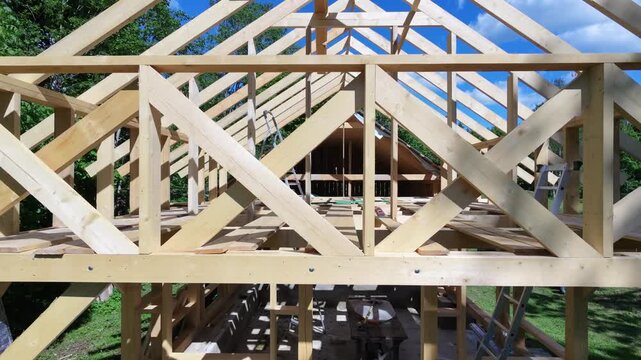 A wooden roof frame rises above an unfinished structure, showing the early stage of building work. Rafters and beams form the outline while tools and loose boards rest on the floor below. 