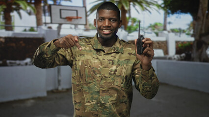 Naklejka premium Soldier in camouflage points finger to handheld radio in building courtyard near basketball hoop and palm trees; pride duty.