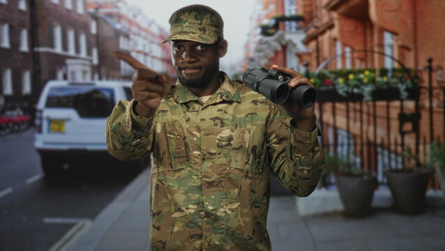 Man soldier in camouflage uniform holding binoculars and pointing finger on a street with parked van and railings; vigilance duty.
