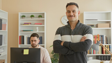 Man standing with arms crossed next to a seated coworker working at a desktop computer amid bookshelves and desk lamp in building  confidence. © Krakenimages.com