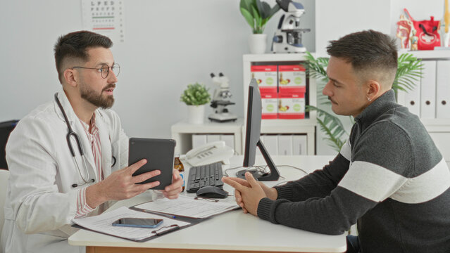 Doctor shows tablet to male patient across desk in medical building while pointing to screen and reviewing notes; calm reassurance.