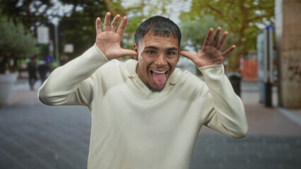 Naklejka premium Young hispanic man sticks out tongue and raises hands to temples in a goofy facial gesture on street, smiling at camera in a cream sweater and trimmed goatee; playful joy.