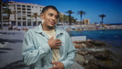 Fototapeta premium Man with hand on chest in a light jacket by palm trees and buildings on a street, smiling and leaning forward; sincere gratitude.