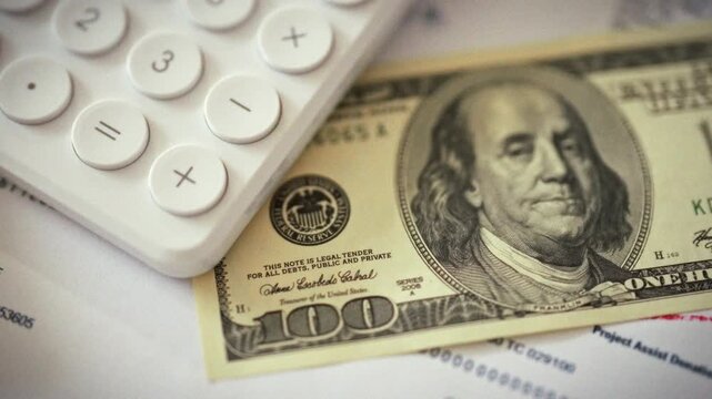Close-up shows crisp paper one-hundred-dollar bill resting beside white calculator, highlighting modern finance. The scene underscores how cash and digital calculation intertwine in money management
