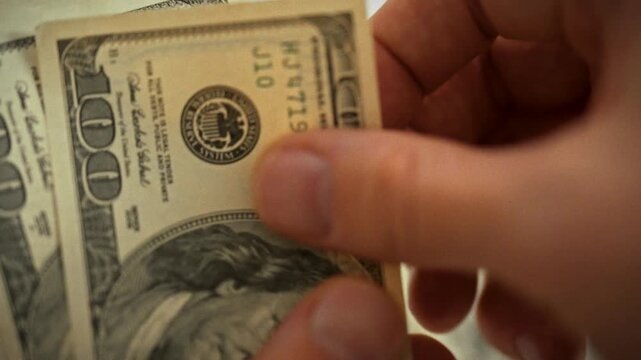 Counting old-style hundred-dollar bills, hands meticulously tally cash, each bill slipping between fingertips as the money is precisely measured, focused routine that underscores financial counting